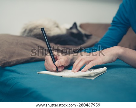 A young woman is lying in a bed with a cat and is taking notes with a pen and notepad