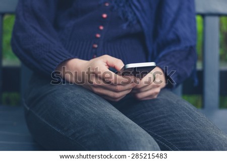 A young woman is sitting on a bench outside in nature and is using a smart phone