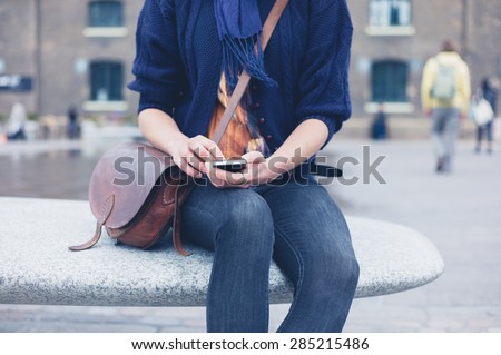 A young woman is sitting outside on a granite bench in the city and is using a smart phone