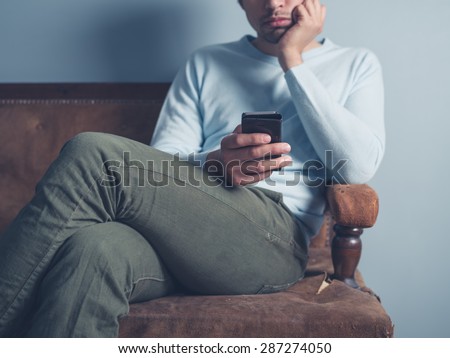 A young man is sitting on an old antique sofa and is using a smart phone