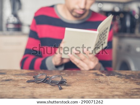 A young man is reading a book in a kitchen. Focus on his reading glasses on the table in front of him.