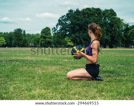 A fit and athletic young woman is sitting on the grass in a park and holding a medicine ball