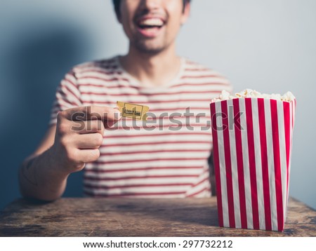 A happy young man is sitting at a table with a box of popcorn and a movie ticket in his hand