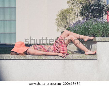 A young woman wearing a dress and a hat is relaxing in a green space outside an apartment building on a sunny summer day