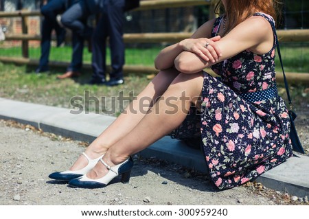 A young woman wearing a dress is sitting outside on a farm with smartly dressed people participating in a social event or gathering