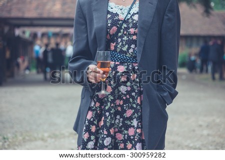 A young woman wearing a man's jacket over her dress is standing with a drink at a party in the countryside