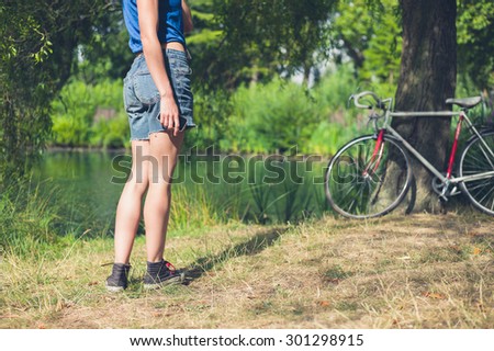 A young woman is standing by the water in a park with her bicycle resting against a tree in the background, she is holding a smart phone in her hand