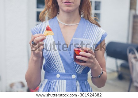 A young woman is standing in a garden in the summer with a drink and a snack in her hands