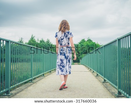 A young woman wearing a dress is crossing a footbridge in a rural setting