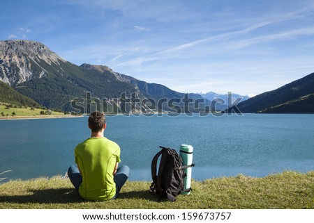 Hiker with backpack and rolled foam camping mattress looking at the lake with mountain background