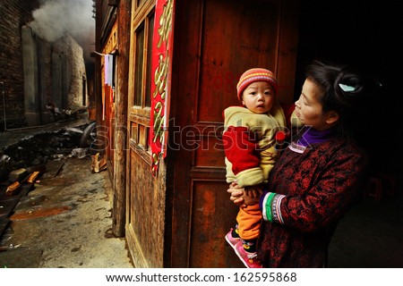 GUIZHOU PROVINCE, CHINA - APRIL 13:  Chinese woman with child in her arms, stands on village street, near wooden house, April 13, 2010. Asian family from rural areas, mother holding son in her arms.