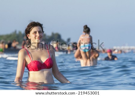 Caucasian teen girl in a pink bathing suit, standing waist-deep in sea water, against many vacationers people who bathe in the sea.