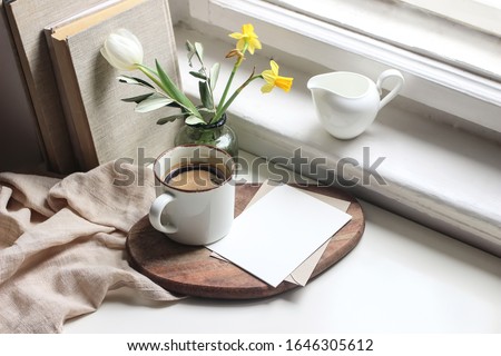 Cozy Easter spring still life. Greeting card mockup scene. Cup of coffee, books, wooden cutting board, milk pitcher and vase of flowers on windowsill. Floral composition. Yellow daffodils and tulip. 