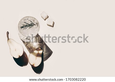Summer stationery still life scene. Glasses of water, rosemary herb, cut pears fruit and ice cubes. Beige table background in sunlight. Vacation refreshment concept. Long harsh shadows. Flat lay, top.