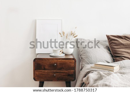 White frame mockup on retro wooden bedside table. Modern white ceramic vase with dry Lagurus ovatus grass and cup of coffee. Beige linen and velvet pillows in bedroom, Scandinavian interior.
