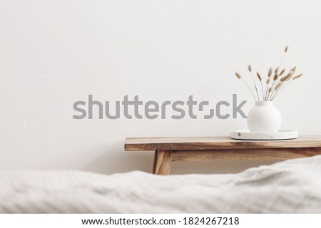 Modern white ceramic vase with dry Lagurus ovatus grass and marble tray on vintage wooden bench, table. Blurred beige linen blanket in front. Scandinavian interior. Empty white wall, copy space.