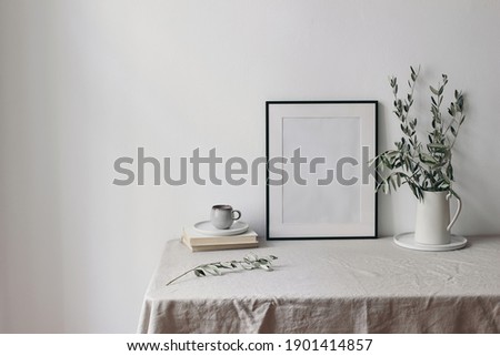 Spring, summer breakfast still life scene. Cup of coffee, books and empty black picture frame mockup. Beige linen tablecloth. Olive tree branches in ceramic jug. Farmhouse, Scandinavian interior.