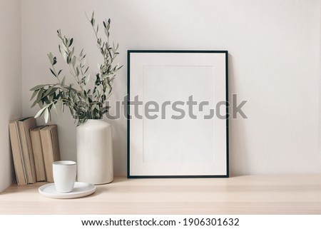 Breakfast still life. Cup of coffee, books and empty picture frame mockup on wooden desk, table. Vase with olive branches. Elegant working space, home office concept. Scandinavian interior design. 商業照片 © 