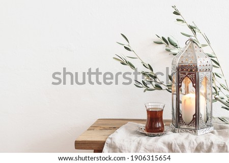 Ramadan Kareem greeting card, invitation. Silver lantern with burning candle. Turkish tea in glass on saucer. Green olive tree branches on old wooden table background. Muslim Iftar dinner. Front view.