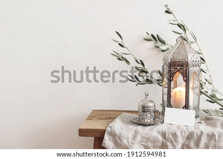 Ramadan Kareem greeting card, invitation mockup. Silver lantern with burning candle and silver cup with tea or coffee. Green olive tree branches on old wooden table background. Muslim Iftar dinner. 