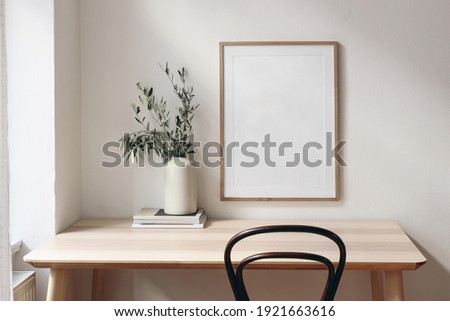 Home office concept. Old books, empty vertical wooden picture frame mockup hanging on white wall. Wooden desk, table. Vase with olive branches. Elegant working space. Scandinavian interior design.
