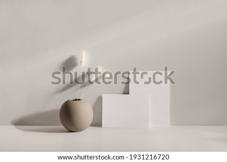 Modern summer stationery still life scene. Beige round vase with dry lagurus grass. Table background in sunlight. Blank business card, invitation mockups lean on champagne wall, long shadows.  商業照片 © 