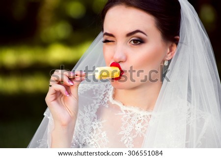 Beautiful bride eating yellow popsicle ice pop looking cute to the side with raised eyebrow on nature background. happy excited expression portrait on nature background. Multicultural Caucasian woman