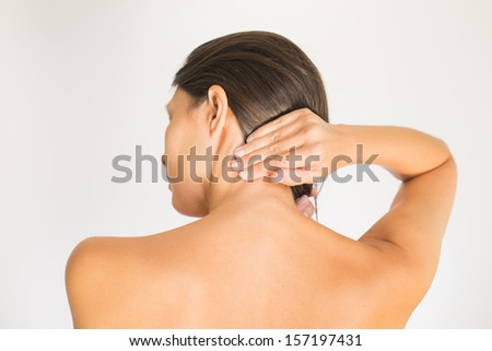 Woman with upper back and neck pain standing naked with her back to the camera and her hand rubbing her shoulder muscles close to the spine