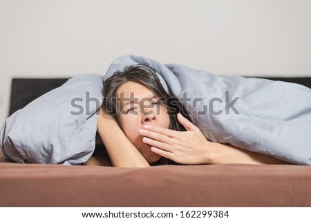 Tired young woman yawning under a duvet holding her hand in front of her mouth as she spends a lazy day relaxing at home