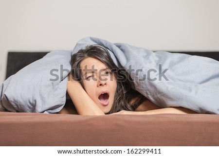 Tired young woman yawning under a duvet as she spends a lazy day relaxing at home