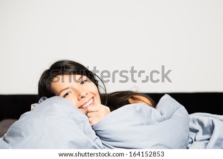 Laughing attractive young woman snuggling up in her comfortable warm bed peeking over the top of the duvet at the camera