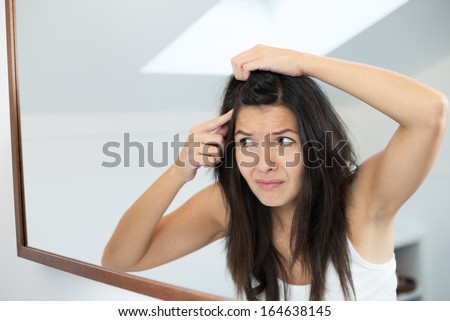 Horrified young woman looking in the bathroom mirror staring open mouthed at the first grey hair on her scalp, a first sign of ag ing, or noticing that she is suffering from dandruff