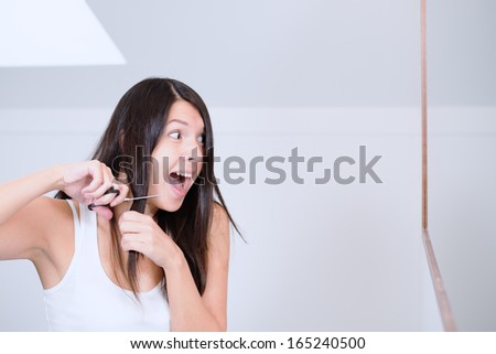 Attractive young woman with long brunette hair preparing to cut it with a pair of scissors in the bathroom grimacing in apprehension as she has misgivings about her decision