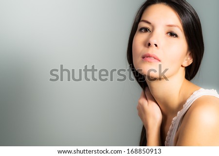 Beautiful young woman with a serious expression standing with her hands to her face looking at the camera in a pensive way, on a gray studio background