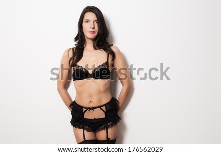 Beautiful woman wearing sexy black underwear with a lacy bra, suspender and stockings standing against a wall with her hand behind her in a tantalizing pose showing off her shapely bare body