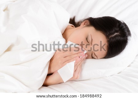 Sick young woman lying in her clean white bed blowing her nose on a tissue conceptual of healthcare and seasonal flu and chills, rhinitis or an allergic reaction in hay fever