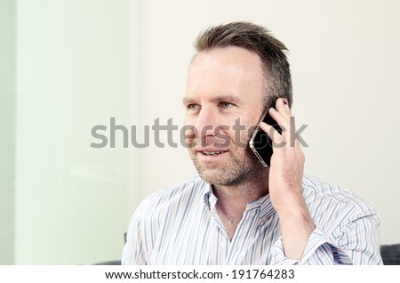 Horizontal portrait of a Caucasian middle-aged handsome man wearing a long-sleeved colorful shirt while having a pleasant conversation on the mobile phone, indoors