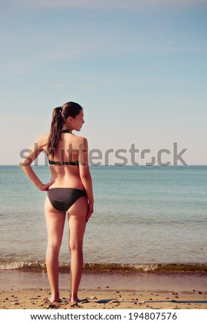 Shapely slender young woman in a bikini standing on the beach with her back to the camera in the summer sunshine facing the calm ocean looking away to her right towards empty copy space