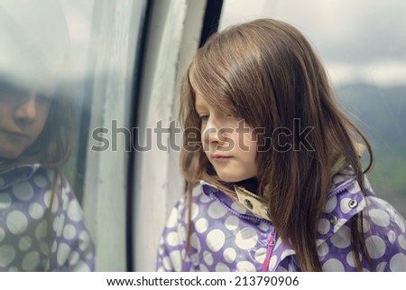 Sad beautiful little girl leaning against a window standing thinking with downcast eyes reflected in the glass