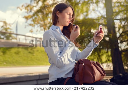 Attractive young woman refreshing her makeup in the street applying lipstick with the use of a small handheld mirror from her large handbag