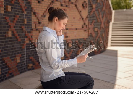 Woman doing business in a commercial foyer sitting on a wall mounted bench against patterned brickwork talking on her mobile phone discussing her notes with a tablet alongside, conceptual of mobility