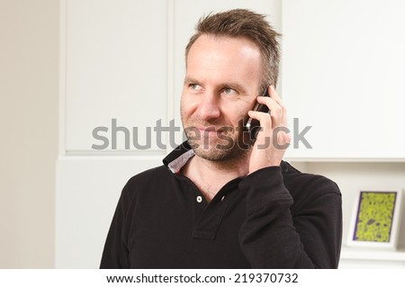 Horizontal portrait of a Caucasian middle-aged handsome man wearing a long-sleeved black T-shirt while having a pleasant conversation on the mobile phone, indoors