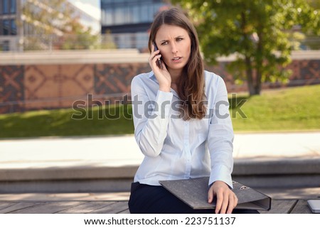 Irritated young woman frowning in annoyance as she sits outdoors in the shade of a tree in an urban park chatting on her mobile phone