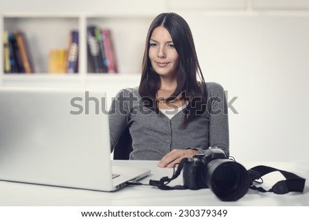 Attractive female photographer sitting back in her chair smiling in satisfaction at her captures as she looks at the screen of her laptop with her camera nearby