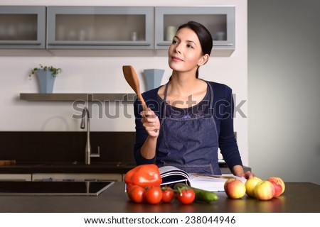 Happy Pretty Woman Wearing Apron Holding Wooden Ladle While Reading a Recipe Book at the Table with Fruits and Veggies in the Kitchen.