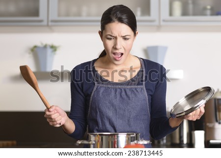 Young housewife having a calamity in the kitchen reacting in shock and horror as she lifts the lid on the saucepan on the stove to view the contents as she cooks dinner