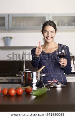 Smiling housewife giving a thumbs up gesture as she enjoys a glass of red wine while standing at the stove cooking dinner from fresh ingredients