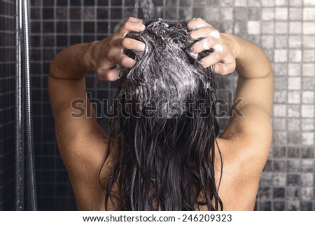 Woman shampooing her long brown hair under a shower standing with her back to the camera working up a lather under the spray of water