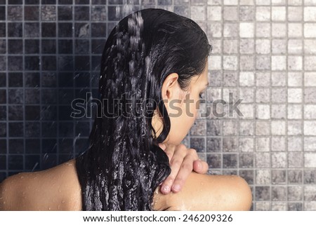 Young woman washing her long hair under the shower standing with her back to the camera rinsing it off under the jet of water with her head partially turned to the side, over grey mosaic tiles