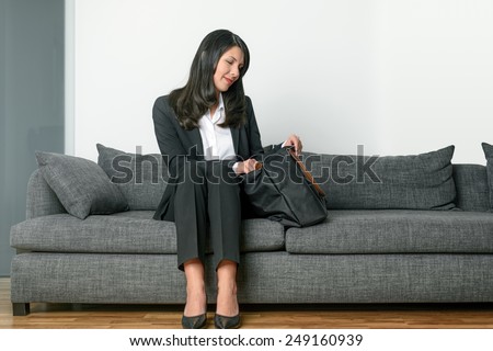 Attractive elegant businesswoman sitting on a grey couch searching through a black briefcase or laptop bag with a smile on her face against a white wall with copy space